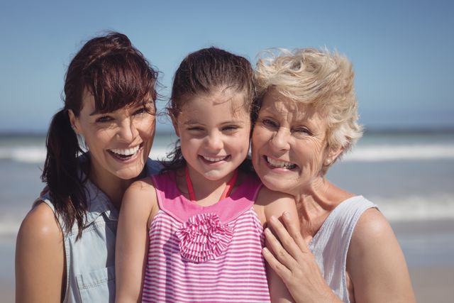 Three generations of women, including a grandmother, mother, and daughter, are smiling and enjoying a sunny day at the beach. This image can be used for promoting family vacations, summer activities, and outdoor fun. It is ideal for advertisements, travel brochures, and family-oriented content.