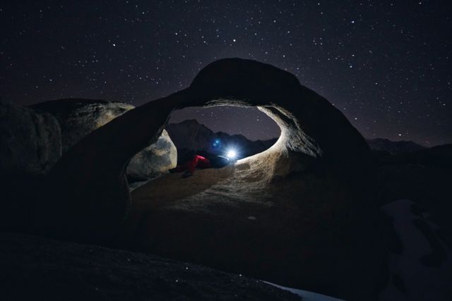 Person exploring natural rock arch at night with flashlight highlighting the formation. Starry night sky creates a captivating backdrop. Ideal for themes related to adventure, outdoor activities, stargazing, astronomy, and night-time expedition.