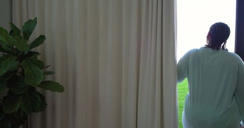 African american woman standing at window drawing curtain, backlit interior, green lawn view