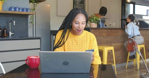 Young woman working on laptop checking phone in bright modern cafe with red coffee mug