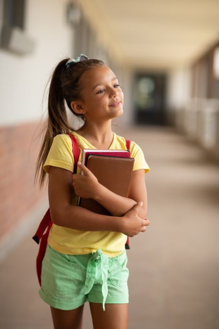 Smiling caucasian elementary schoolgirl with books looking away while standing school corridor - Download Free Stock Photos Pikwizard.com
