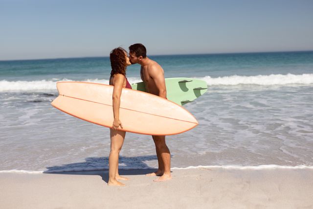 Romantic couple kissing on beach while holding surfboards. Perfect for travel brochures, summer vacation promotions, relationship blogs, and advertisements for beachwear or surfing equipment.