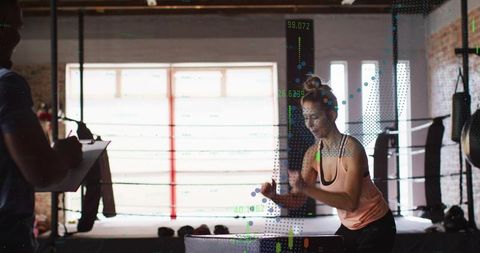 Female athlete performing explosive box jump in boxing ring while trainer recording metrics