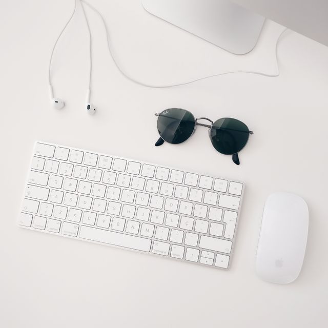 Minimalist workspace showing white keyboard, mouse, round sunglasses, and earphones on a white desk. Perfect for blog posts on minimalism, technology setup ideas, or workspace organization tips. Ideal for presentations or websites focusing on tech accessories and modern office aesthetics.