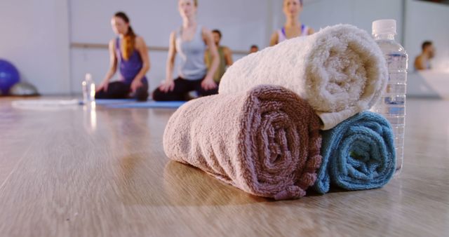 Rolled towels and a water bottle sitting on a wooden floor in a gym or yoga studio while women are seated in the background post-exercise. Ideal for promoting fitness classes, yoga sessions, or healthy lifestyle products, emphasizing the importance of hydration and relaxation post-workout.