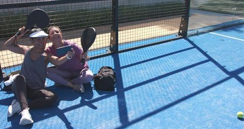 Two women taking selfie on blue padel court holding paddles and phone, smiling in sunlight