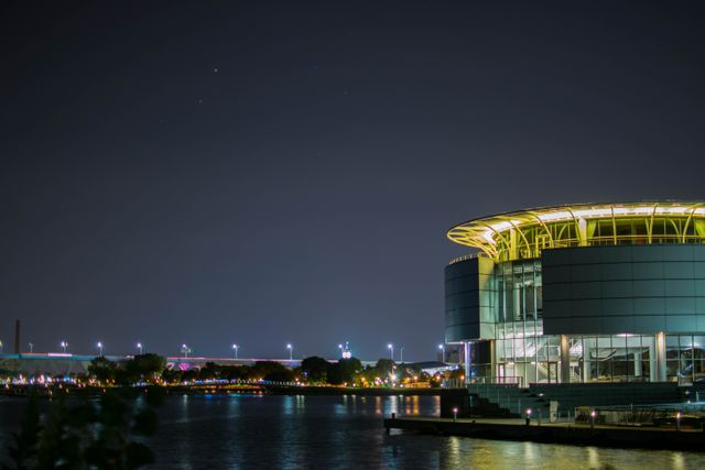 This scene features a modern building illuminated by lights, situated by a riverside with a clear night sky in the background. The reflections on the water add a serene charm to the evening urban landscape. This visual would be perfect for content related to modern architecture, urban living, nighttime photography, or waterfront cityscapes.