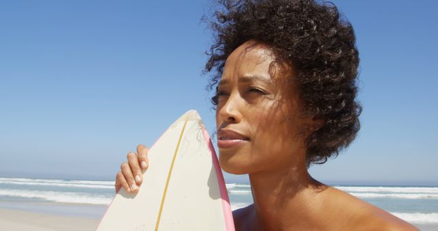 African American woman holding a surfboard on a sunny beach day, with copy space. Her contemplative gaze suggests she's preparing for a surf session or reflecting on her day at the sea.