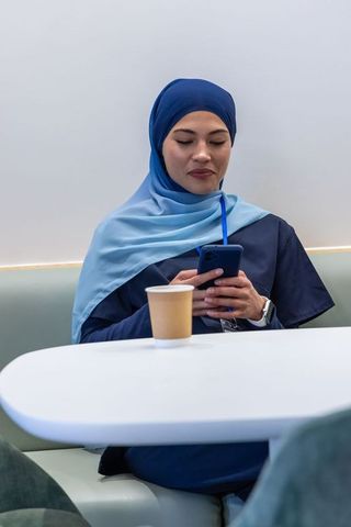 Woman in blue headscarf scrolling smartphone during break in modern cafeteria