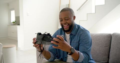 African american man laughing holding sneaker on sofa in bright modern living room