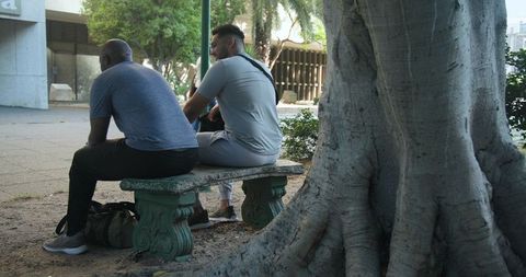 Two african american men sitting on stone bench in urban courtyard under large tree