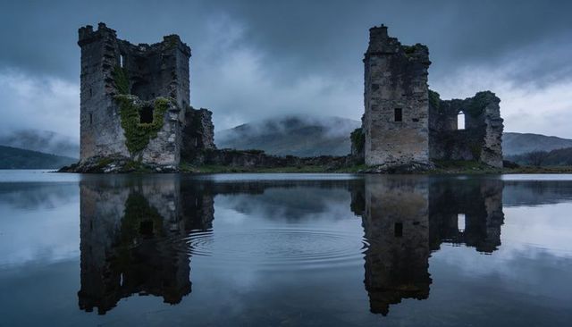 Misty lochside castle ruins looming and reflecting in calm water with concentric ripples