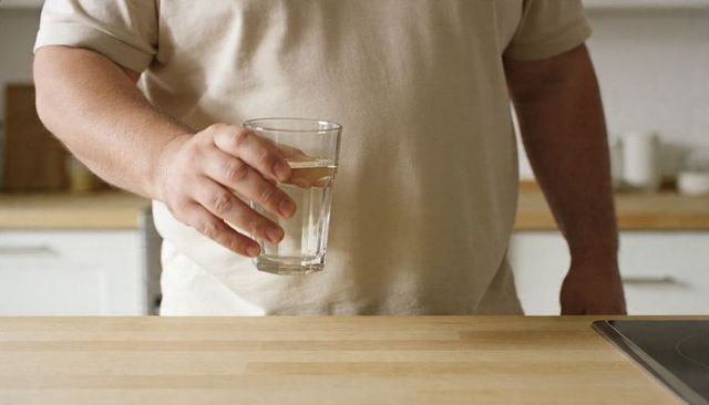 Man holding glass of water at light wood kitchen counter wearing beige t-shirt