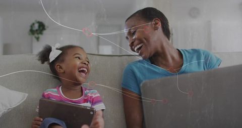 Laughing mother and daughter connecting on sofa with tablet and laptop for home bonding