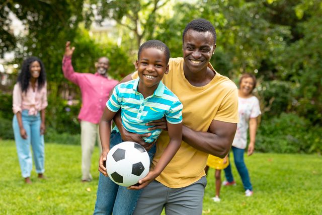 Cheerful african american man carrying son holding soccer ball while family in backyard. unaltered, leisure, playful, fun, togetherness, weekend activities, sport.