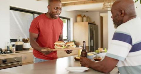 Young man serving burgers to father at sunlit kitchen island, casual family lunch moment