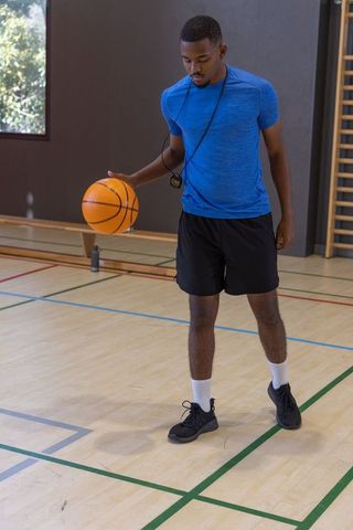 African american coach dribbling basketball while using stopwatch during indoor training