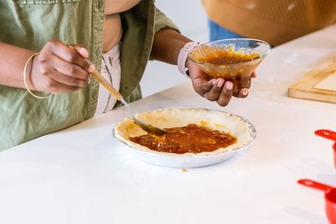 African american woman spreading jam into pie crust on white countertop for homemade baking