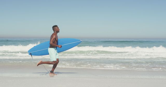 Young male surfer holding blue surfboard running on sandy beach along the shoreline with ocean waves in the background. Perfect for promoting summer activities, beach lifestyle, surfing lessons, athleticism, travel and tourism.