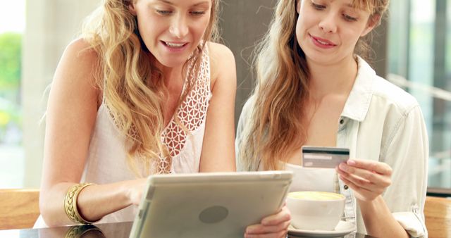 Two young Caucasian women are engaged in online shopping or financial transactions using a tablet and a credit card. Their focused expressions and the casual setting suggest a blend of leisure and commerce, indicating a moment of shared decision-making or purchase.