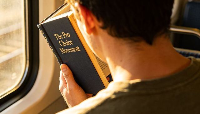 Commuter reading and holding book titled pro choice movement on sunlit train at golden hour