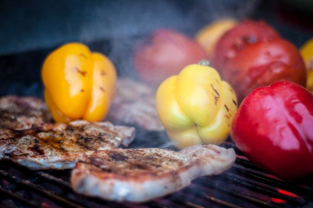 Steaks and colorful bell peppers are grilling on an outdoor barbecue. Smoke and heat enhance the flavors of the food. Suitable for use in articles or advertisements about summer barbecues, outdoor cooking, healthy eating, grilling techniques, or recipes involving grilled vegetables and meat.