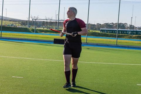 Field hockey player standing on turf holding red stick, looking left, wearing maroon socks