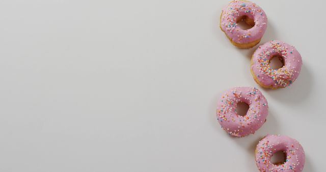 Four donuts covered in pink icing and colorful sprinkles arranged on plain white surface. Useful for advertisements related to bakeries, cafes, confectioneries, and dessert promotions. Perfect for social media posts, blog articles, and recipe websites focused on sweet treats and desserts.