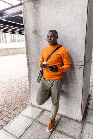 African american commuter leaning on concrete pillar wearing orange sweater holding phone