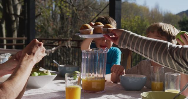 Family gathered outdoors enjoying breakfast on a sunny morning. Muffins are being served along with glasses of orange juice, indicating a warm and inviting environment. Trees and a sunlit view are visible in the background. Perfect for advertising family gatherings, healthy eating, and outdoor activities.
