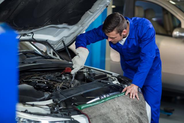 Mechanic in blue uniform and gloves repairing car engine in automotive workshop. Useful for illustrating automotive repair, car maintenance, mechanical services, and professional technicians at work.