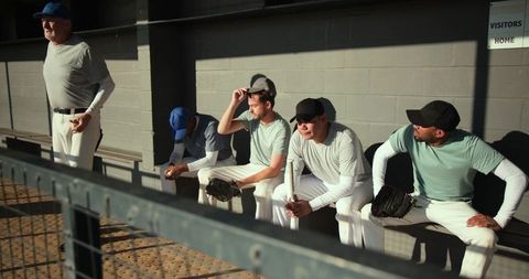 Baseball teammates sitting in dugout waiting for game, holding gloves and bat in sunlight