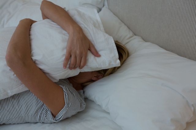 Woman lying in bed covering her face with a pillow, creating a sense of comfort and relaxation. Ideal for use in articles or advertisements related to sleep, relaxation, home comfort, and bedroom decor.