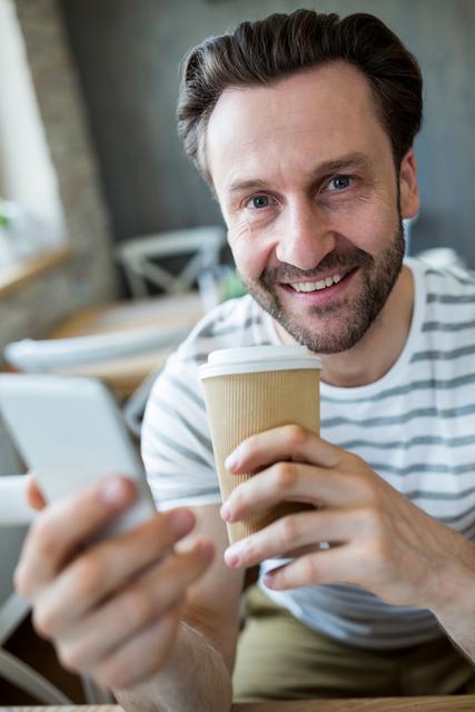 Portrait of smiling man holding mobile phone and disposable coffee cup in coffee shop