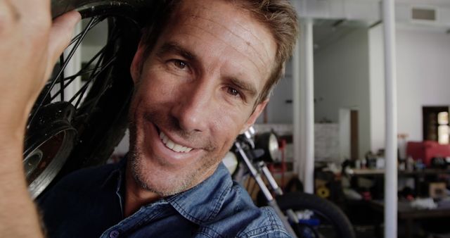 Middle-aged mechanic smiling while carrying a bicycle wheel in a workshop filled with tools. Ideal for content related to bicycle repair, craftsmanship, or do-it-yourself projects. Suitable for promoting mechanical workshops, services, or instructional materials.