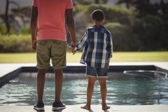 Father and son standing together by the poolside on a sunny day, holding hands and enjoying quality time. Perfect for use in family-oriented advertisements, parenting blogs, summer vacation promotions, and outdoor lifestyle content.