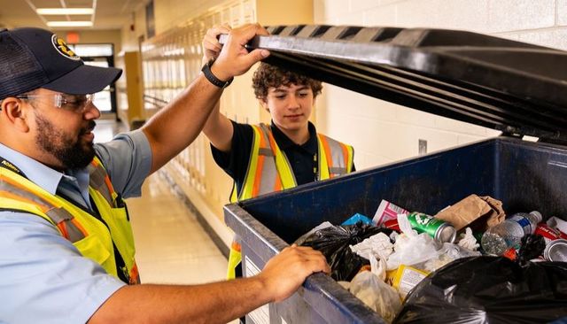 Custodian teaching student recycling while lifting dumpster lid in school corridor