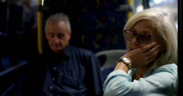 Senior couple traveling by bus at night, with the woman resting her head on her hand and wearing glasses. Ideal for depicting themes of aging, travel, public transportation, and the quiet moments during daily commuting. Great for articles about elderly lifestyles, public transportation use among seniors, or serene travel moments.