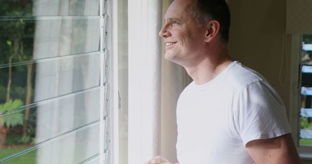 This image of a smiling man in a white t-shirt looking out the window is perfect for promoting concepts of positivity, relaxation, and overall well-being. It can be used in lifestyle blogs, mental health articles, advertisements for home or interior brands, or any content emphasizing personal happiness and daily comfort.