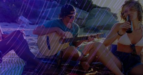 Friends playing acoustic guitar and drinking from bottles on sunlit rocky beach at sunset