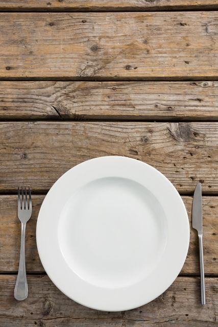 Overhead view of a simple dining arrangement with an empty white plate and stainless steel cutlery on a rustic wooden table. Ideal for use in food blogs, restaurant websites, and home decor magazines to illustrate topics related to dining, meal preparation, or minimalist table settings.
