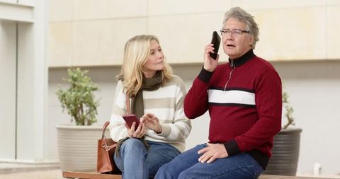Senior couple sitting on bench, man talking on phone while woman checking smartphone