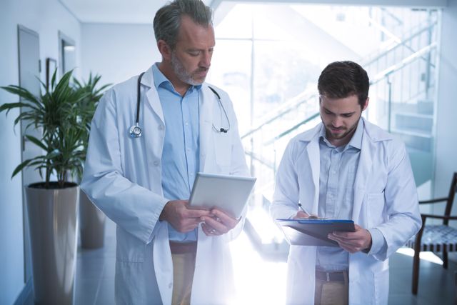 Two doctors are collaborating in a hospital corridor. One is using a digital tablet while the other is writing on a clipboard. This image can be used for healthcare-related content, medical teamwork, modern medicine, and hospital environments.