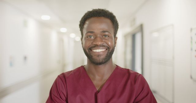 Male healthcare professional smiling in hospital corridor, wearing red scrubs. Image can be used for promoting healthcare services, medical staff recruitment, healthcare awareness campaigns, and hospital brochures. Emphasis on friendliness and professionalism in healthcare settings.