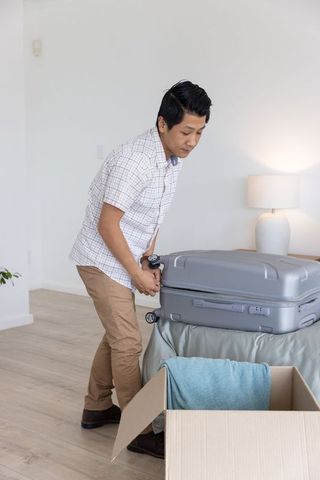 Middle-aged asian man packing suitcase on bed during move in bright modern apartment