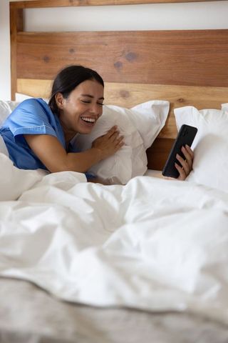 Laughing woman in blue pajamas using smartphone on white bed for cozy morning self-care