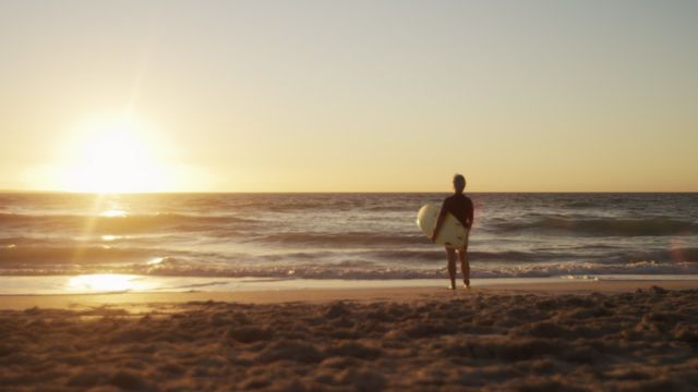 Senior man holding surfboard standing on sandy beach facing ocean at sunset. Suitable for themes like retirement lifestyle, relaxation, surf culture, beach holidays, and enjoying life moments.