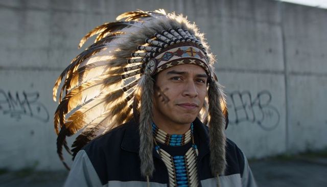 Standing man wearing feathered warbonnet and beaded turquoise choker against graffiti wall