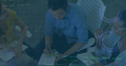 Collaborative team leaning over notebooks and writing notes while financial charts overlay