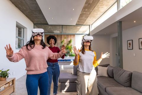 Diverse women playing virtual reality in open-plan living room, laughing and reaching